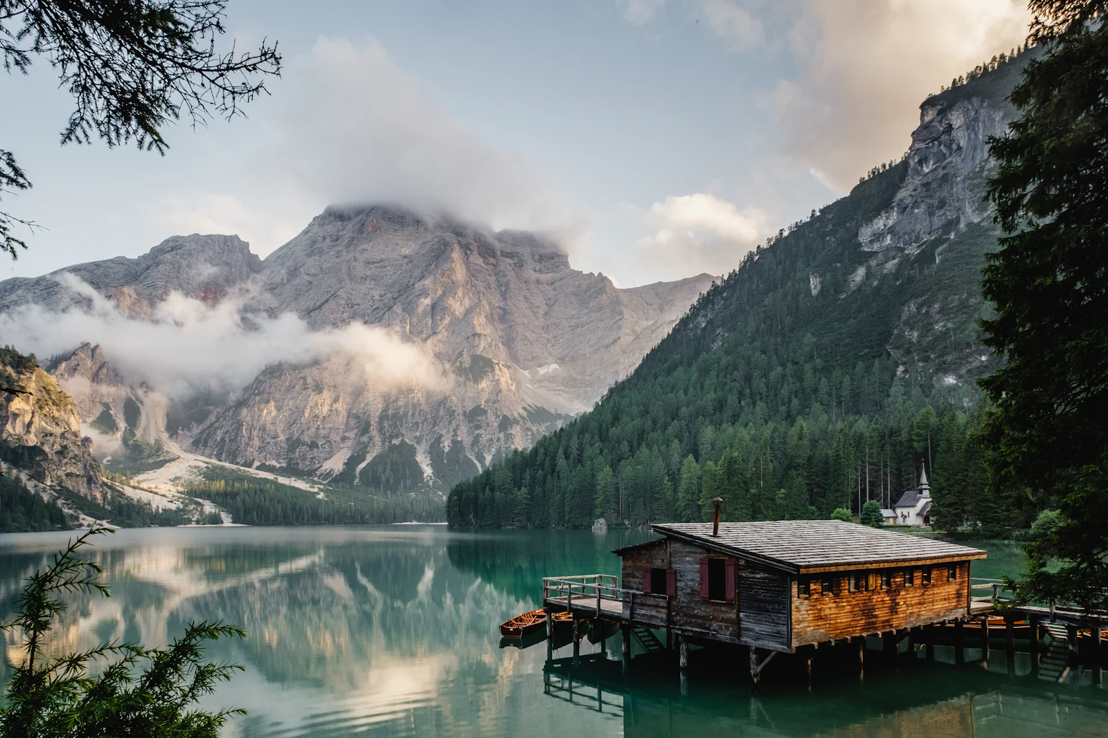 A still alpine lake beneath the Dolomites, with a small wooden boathouse at the water’s edge.
